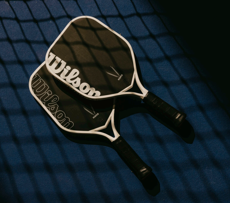 Two Wilson pickleball paddles laying on a court surface with the net casting its shadow over them